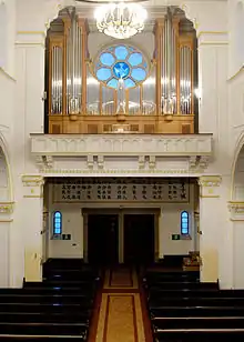 Interior of the end of the cathedral showing the main door with a choir loft above it on which stands a large pipe organ above which is a blue rose window with a dove at its centre