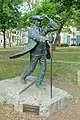 A bronze statue situation in a park surrounded by trees showing a standing man wearing a tricorne hat and frock coat. The man is holding the hat on his head with his left hand and has a walking pole in his right hand. His coat is billowing out behind him.