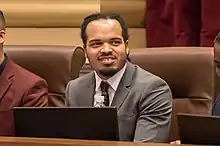 A young, black man with a mustache and anchor beard sits behind a laptop and stares off to his left. Another person, barely visible, is seated to Ellison's right.