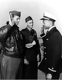 Three man stand on the deck of a ship, one with his arm raised and another reading from a paper.