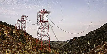 Towers supporting one of two antenna feed bus cables extending from the transmitter building.  From each 145 ft. tower a vertical feedline (faintly visible, top of picture) extends up 900 ft. to attach to the midpoint of each horizontal valleyspan cable.