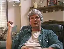 A casually dressed, middle-aged white man with wavy gray hair sits in an overstuffed chair in a living room