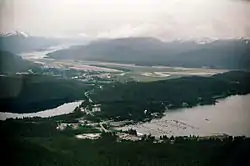 Aerial view shows Auke Bay (including the harbor and Auke Lake) in the foreground. The Mendenhall Peninsula extends to the right behind the community. The lower Mendenhall Valley, Juneau International Airport and Douglas Island are in the background.