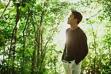Justin Nozuka wearing light pants and a dark long-sleeved shirt, standing in profile within a natural landscape