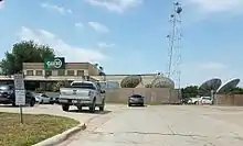 A low-slung, tan industrial building with a green CW 33 sign. Satellite dishes and a communications tower are visible on the side of the building.