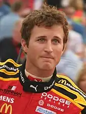 A man in his late twenties wearing black sunglasses with a head full of hair and a red jacket with sponsors' logos.