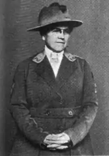 Middle-aged white woman, wearing glasses and a uniform-style jacket and hat, with hands folded in front of her body