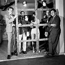 Black and white photograph of Kenneth Kemble, Lopez Anaya, Silvia Torras, Luis Wells, and Jorge Roiger. The middle three people are standing behind a large frame with mannequin heads hanging from it.