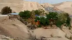 The image shows buildings amongst green trees, with solar panels at the top, surrounded by sandy terrain. A highway with several cars is seen in the background.