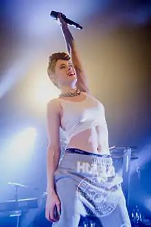 Kiesza wearing a white crop top and grey sweatpants, standing onstage with one arm raised, holding microphone