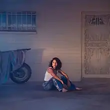 A photo of the artist sat on the ground in front of a house, wearing a white tank top and blue jeans and staring off to the side at the middle distance. Behind her is the wall of the house, dimly lit, with an ornate metal door to the right and a window with metal bars over it to the left with a motorcycle covered in a tarp leaning against the wall below it.