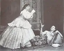 woman kneeling in front of a standing man; the two are conversing and each is gesturing with one hand as if ringing a small bell