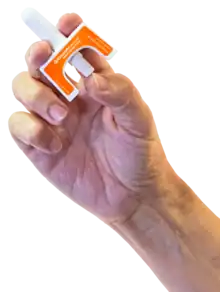 A hand holding a Kloxxado intranasal spray device. With the tip inserted into the nostril of someone who has suspected overdose, a dose of naloxone is administered by using one's thumb to push the plunger up toward the index and middle fingers.