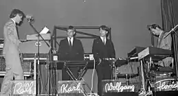 A black and white photograph of four members of Kraftwerk onstage, each with a synthesizer