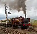A pannier tank locomotive is passing a semaphore signal, leading a train which includes one visible passenger carriage. The pannier tanker is maroon, apart from the black chimney, brass safety valve cover,  red buffer beam, and grey coupling rods. The tank, cab, splashers and toolbox are all lined in yellow. London Transport is written on the side of the tank, and L.94 on the side of the cab, again both in yellow. A plume of brown smoke comes from the chimney, and there is as whisp of steam from under the pannier tank. The passenger carriage is cream and brown with a grey roof.