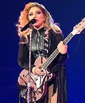 Lady Gaga standing behind a microphone stand with a pink guitar in her hands, wearing black leather fringe.