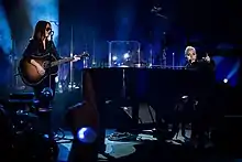 Gaga playing piano and Hilary Lindsey standing opposite to her paying guitar onstage.