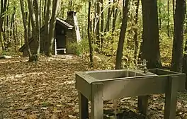 A metal sink with running water supported by wooden beams in the foreground, a wooden open shelter with a stone chimney amidst the trees in the background