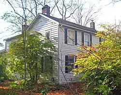 A white house with black shutters and brick chimneys seen from its front left corner. Shrubs and trees obscure the view on the sides.