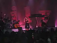 A woman singing on a microphone, standing up, with a band with instruments in her back and a crowd of people in front. There are 3D letters in the background, spelling "Lily".