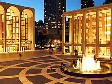 The corner of a lit up plaza with a fountain in the center and the ends of two brightly lit buildings with tall arches on the square.