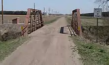 Bridge on dirt road with fields in distance; railcars at right edge of picture