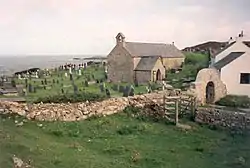 A small windowless church in a churchyard, with the sea visible in the distance. A stone arch is visible over the path leading into the churchyard.