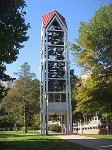 A three-dimensional vertical structure perhaps 70 feet (21 m) tall and 10 feet (3.0 m) wide by 10 feet (3.0 m) long is filled with tiers of large metal bells. The structure, standing in front of a large institutional building in a park-like setting, is supported by several metal legs. A sidewalk passes through the structure, which is open at the bottom to a height of perhaps 10 feet (3.0 m).
