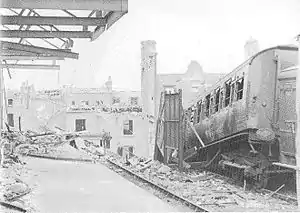 A railway platform strewn with rubble. Broken girders jut out above the platform, while alongside the platform a set of railway lines stop abruptly at a large crater. Next to the railway line is a burned-out railway carriage tilted at an acute angle.