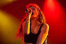 A picture of Lorde performing at a concert. Her face is captured singing in a dramatic pose, wearing a black crop top with yellow and red lights illuminating the stage.