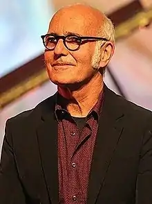 balding, white-haired Caucasian male wearing glasses and a black jacket, smiling and looking right of camera