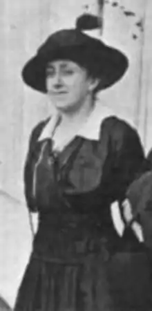 A white woman, standing outdoors, wearing a dark dress with a white collar, and dark brimmed hat