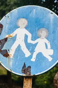 A road sign depicting an adult walking across the stret holding the hand of a child, who is carrying a lunchbox. The colors are white on blue and the sign is faded and chipped.