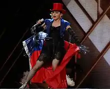 Madonna in a red hat and blue jacket with a red-and-blue flag wrapped around her performing onstage