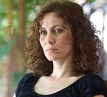 Head shot of Maha Haj, she has shoulder-length curly brown hair. The background is blurry trees