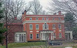 A large two-story flat-roofed rectangular brick building with four corner chimneys classically inspired ornamentation. There is a large full-height wing on the left and a small white wooden porch on the right. There are bare trees and a car parked in the front driveway.