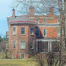 The same house seen from the side with the large wing, also in springtime, illuminated by sun from the left, with some bare branches and bushes partially obscuring the view