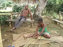 A Malagasy girls sits on the ground weaving with reeds, while an older gentleman sits on a bench above her.