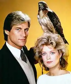 A man in a suit, holding a hawk. Beside him a blonde woman.