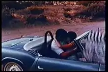 A man and woman kiss in a convertible sports car, with the clapperboard at the edge of the frame.