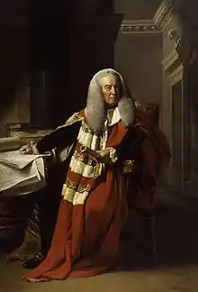 A full-length portrait of an elderly man, seated. He wears long flowing red and white robes, a long grey wig, and holds a rolled document in his left hand. His right hand rests on a table littered with documents. Behind him, the corner of a room, with ornate plaster architrave, is visible.