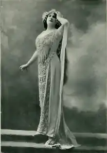 A white woman, standing, hand on forehead, wearing a gown and headdress typical of 1900s Wagnerian opera.