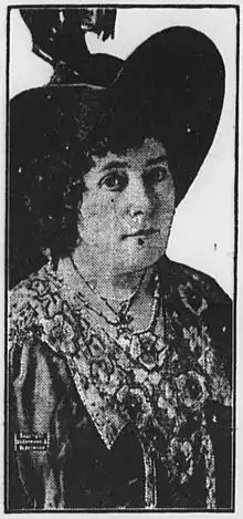 A white woman wearing a large dark brimmed hat and a heavily beaded/embroidered dress