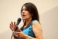 A brunette Caucasian woman wearing a blue blouse is holding a microphone.