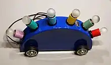 A dark blue half- moon shaped lamp with wheels, decorated with six lightbulbs with colored holders in pink, greeen, blue, yellow and red.