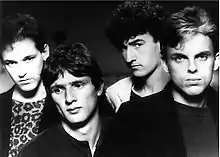 Head and shoulders shot of four men. Man at left has dark hair, face slight tilted to his right, but looking forward, he wears a dark jacket over a print tee-shirt. Second man is the shortest, also dark haired, his head is tilted further with eyes looking to his right, he wears a dark tee-shirt. Third man is the tallest, also dark haired, with his face partly in left profile, his head is also titled, his eyes are direct to the viewer, he wears a light coloured jacket with a dark tee-shirt. The last man is facing forward, he is the second tallest, his hair is lighter with fair coloured streaks at front, his jacket is dark over a less dark tee-shirt.