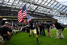An American football player in blue and gold uniform holding a United States flag charges onto the playing field as photographers and teammates cheer.
