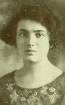A young woman with fair skin and dark hair cut in a wavy bob; she is wearing a dress or top with a scooped neckline and embroidered flower designs