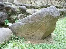 A roughly triangular boulder with the point inclined to the right. It has been carved in low relief to outline the jaws, teeth and eyes of a crocodile, with the point of the boulder representing the snout. A stonework structure rises behind the sculpture, which faces towards a grassy area to the right