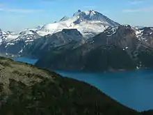A prominent, rocky mountain with heavily glaciated lower slopes rising over mountainous terrain and an alpine lake.